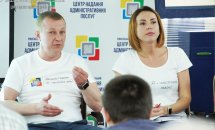 Чоловік та жінка у білому одязі сидять у приміщенні. Чоловік щось розповідає.