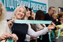 Жінка світлим волоссям тримає стрічку у руках, позаду - інші люди.