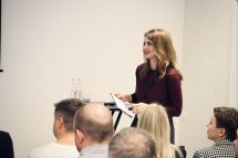 Жінка стоїть перед людьми та тримає в руках аркуші паперу.