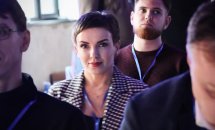 Жінка з коротким волоссям сидить серед чоловіків.