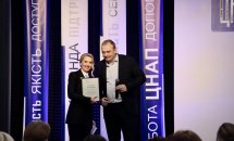 Жінка в чорному костюмі тримає подяку, біля неї чоловік з мікрофоном