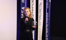 Жінка в чорному одязі тримає в руках мікрофон та подяку