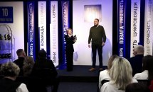 Жінка з чоловіком стоять на сцені, по боках сидять люди