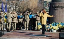 Жінка співає біля пам'ятника, збоку стоять чоловіки у військовій формі.