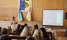 Жінка стоїть біля екрану, в залі сидять багато людей.