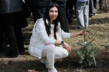 Жінка у білому одязі сидить біля саджанця ялинки.