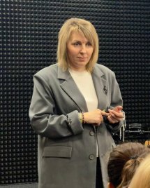 Жінка у сірому піджаку на фоні стіни.