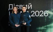 Дві жінки з нагородами позують на фоні банера.