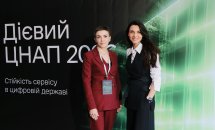 Дві жінки фотографуються на фоні банера.