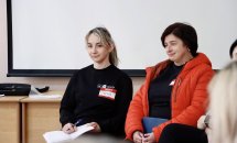 Жінка в чорній кофті та  жінка оранжевій куртці сидять у приміщенні.