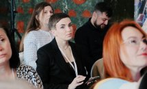 Жінка сидить у приміщенні, навколо неї сидять інші люди.