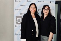Дві жінки в темному одязі позують на фоні банера.
