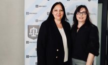 Дві жінки в темному одязі позують на фоні банера.