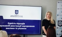 Жінка зі світлим волоссям стоїть біля екрану.