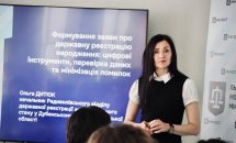 Жінка із темним волоссям стоїть біля екрану.