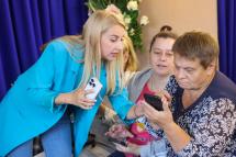 Жінка зі світлим волоссям тримає телефон у руках, та показує щось в іншому телефоні для жінки, яка сидить.