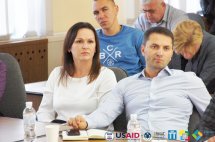 Чоловік та жінка сидять у приміщенні. Позаду сидять інші люди.