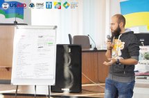 Чоловік з бородою тримає мікрофон та щось розказує перебуваючи біля дошки із записами.