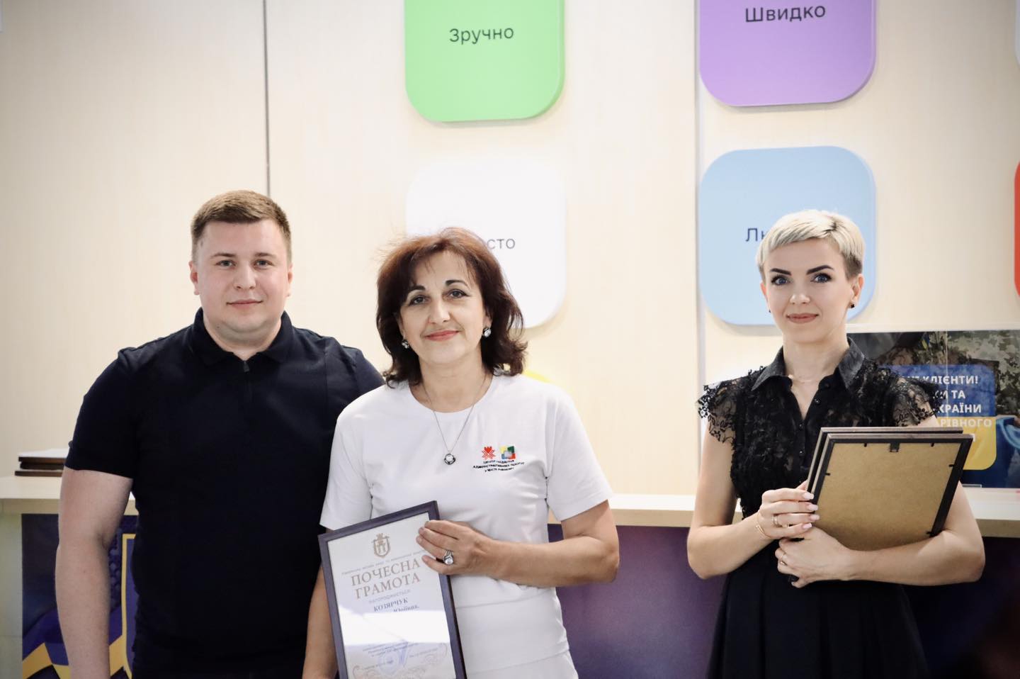 Троє людей стоять разом. Жінка в центрі тримає почесну грамоту. Напис на її футболці: 'Центр надання адміністративних послуг'.