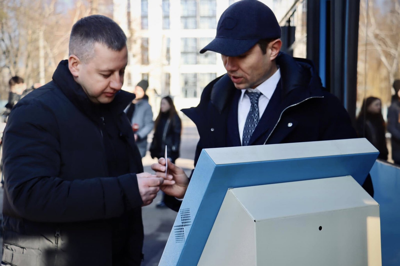 Двоє чоловіків стоять на вулиці біля терміналу. На задньому фоні прохожі та тролейбус.