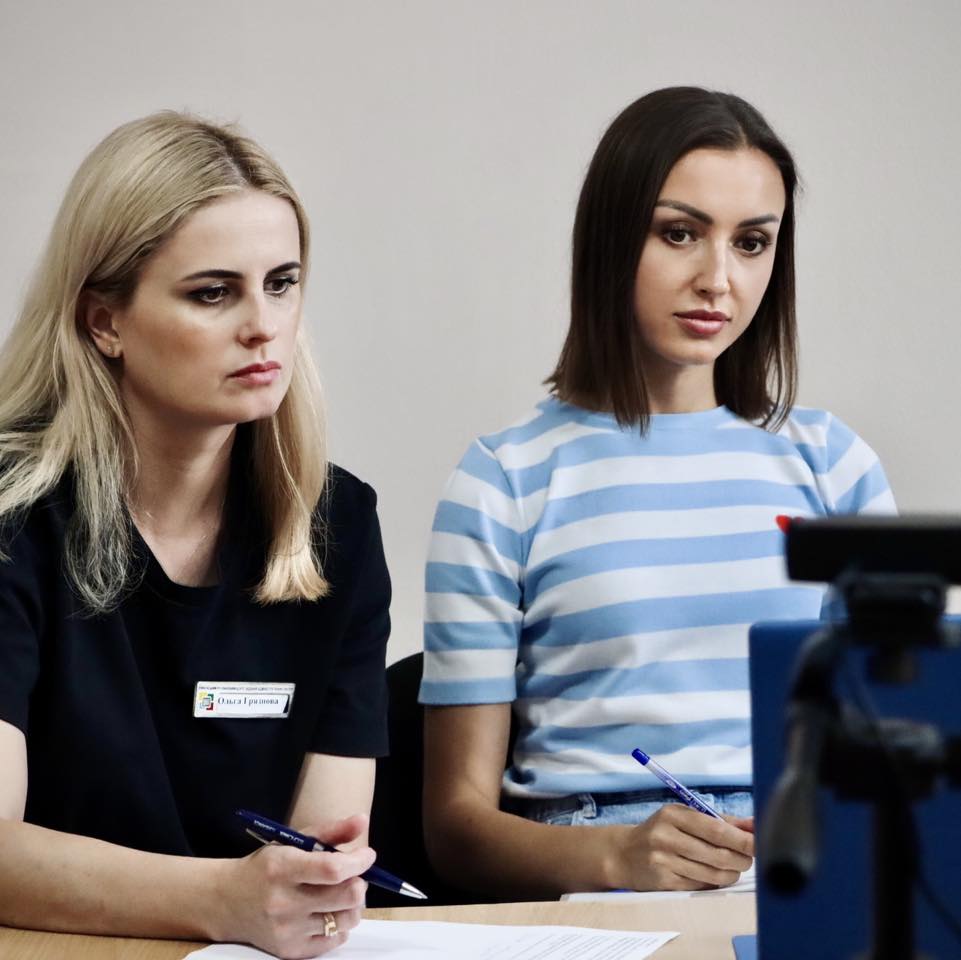 Дві жінки сидять за столом, уважно дивлячись на екран перед собою. Ліва жінка одягнена в чорну сорочку з бейджиком, права - в синьо-білу смугасту сорочку. Обидві тримають ручки та блокноти.