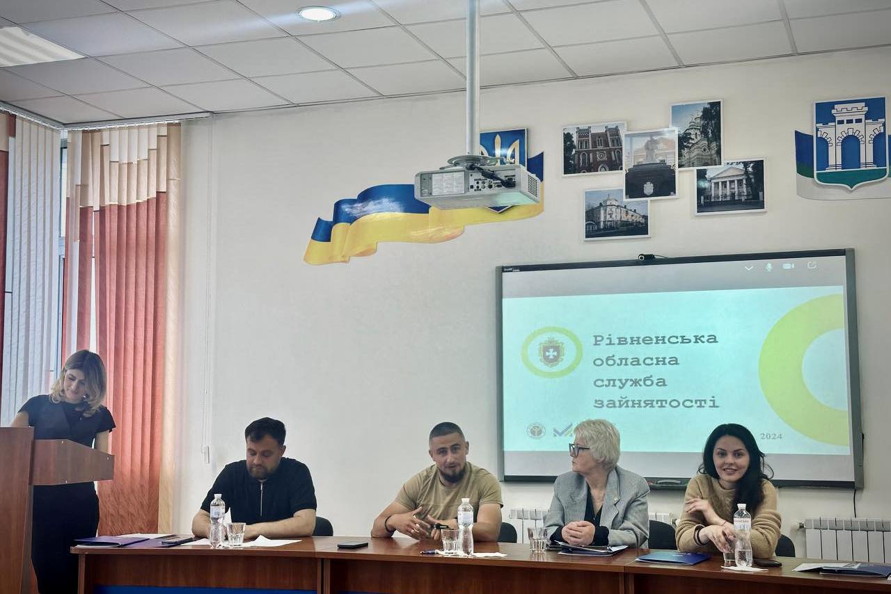 Група людей сидить за столом під час зустрічі у приміщенні Рівненської обласної служби зайнятості. На передньому плані п’ятеро осіб, серед них жінка за трибуною зліва, двоє чоловіків та дві жінки, які сидять за столом. На стіні висить екран з логотипом та назвою служби, а також зображені український прапор і фотографії будівель.