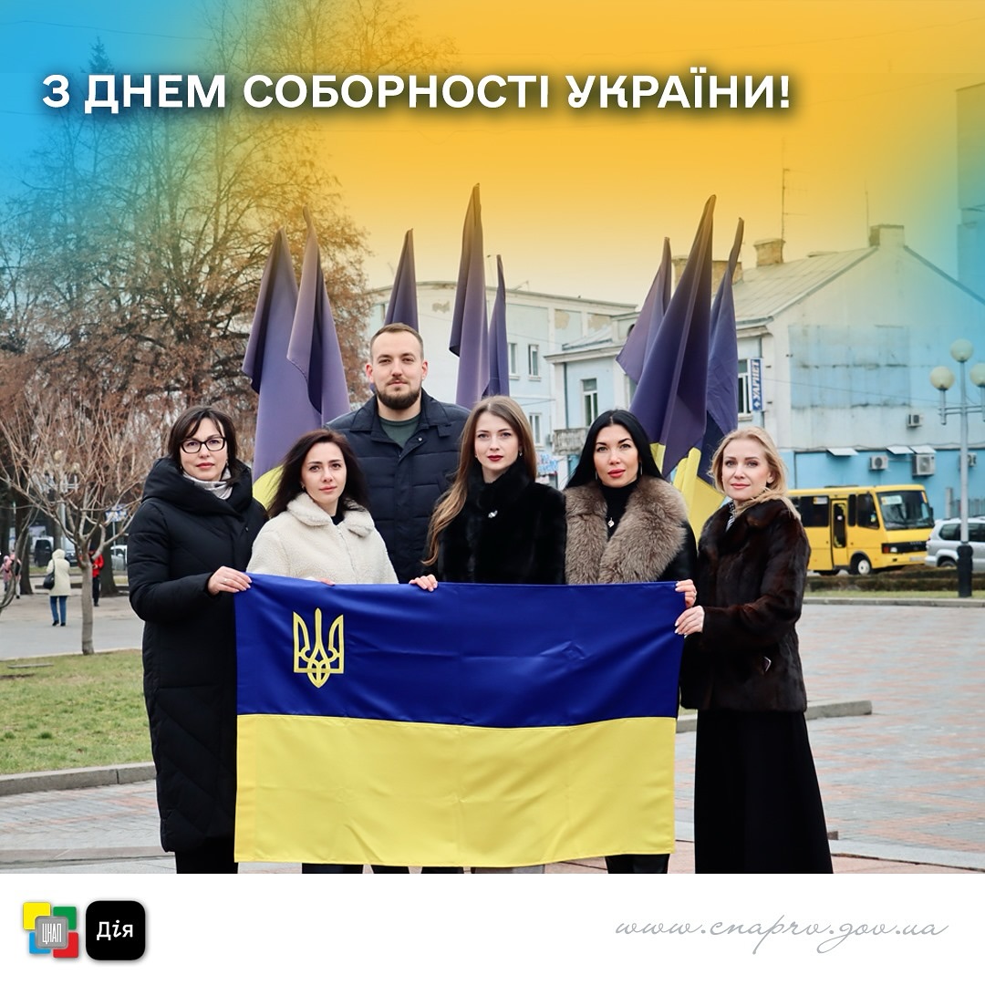 Чоловік та жінки на вулиці тримають прапор України. На задньому плані видно місто Рівне. 