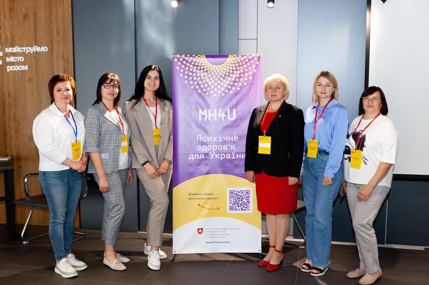 Шестеро жінок стоять біля банера з написом MH4U Психічне здоров'я для України та дивляться в камеру. Вони носять бейджі різного кольору на червоних стрічках. Жінки одягнені в діловий стиль.