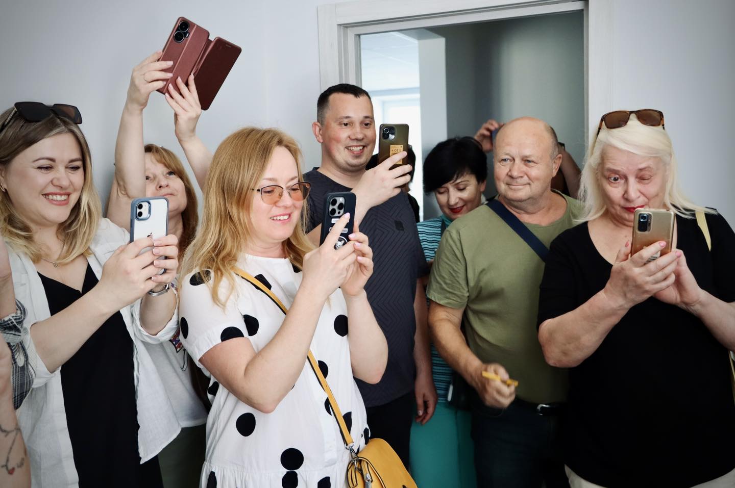 Група людей стоїть разом у кімнаті, усміхаючись і фотографуючи щось на свої смартфони.