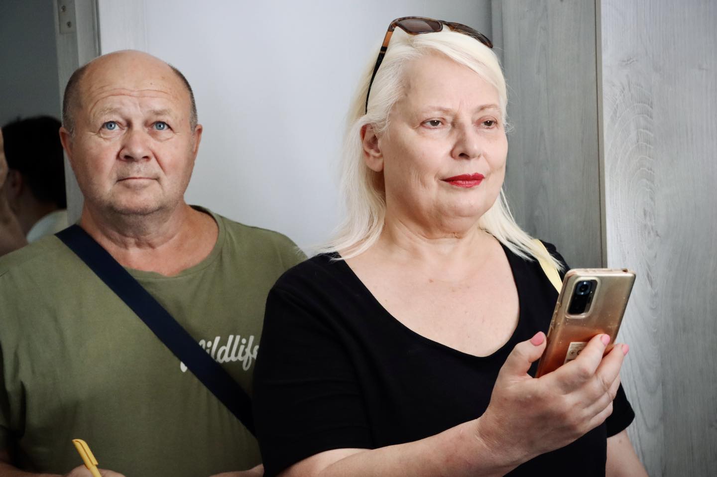 Чоловік і жінка стоять разом; чоловік у зеленій футболці з сумкою через плече, а жінка у чорній блузці з білим волоссям і сонцезахисними окулярами на голові, тримає смартфон.