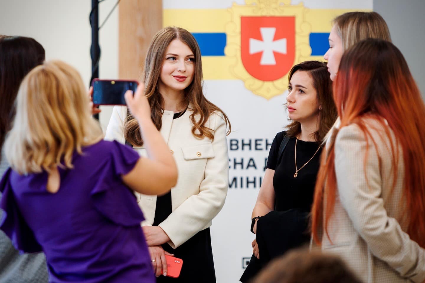 Група жінок стоїть перед екраном із гербом та текстом на тему вебдоступності. Одна жінка знімає на телефон інших, що спілкуються.
