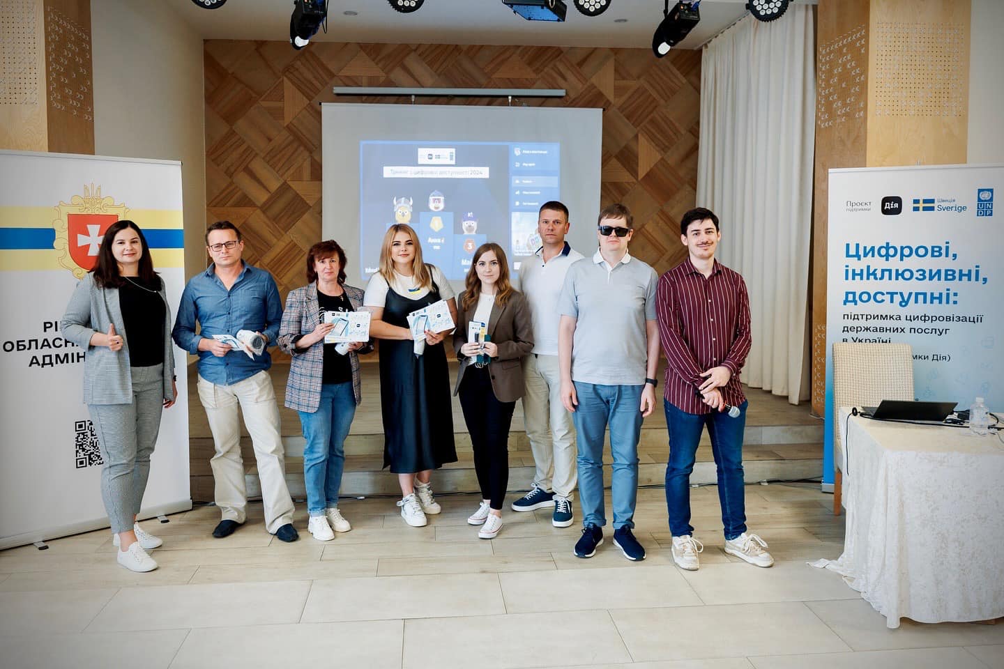 Вісім людей стоять у ряд у приміщенні перед проєктором та банерами. Деякі тримають у руках сертифікати. Позаду них проєктор показує слайд із назвою заходу. Ліворуч і праворуч стоять банери з логотипами та інформацією про підтримку цифровізації державних послуг в Україні.