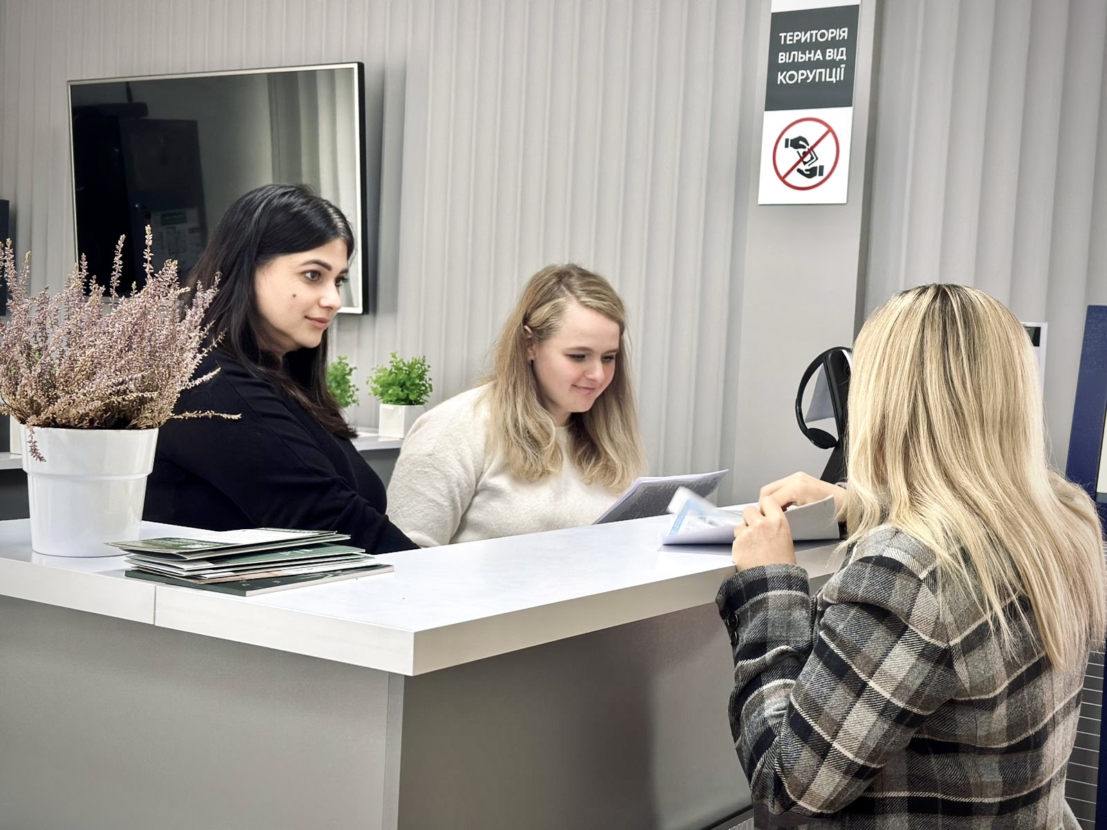 Три жінки розмовляють біля стійки у приміщенні, на стіні позаду висить телевізор.