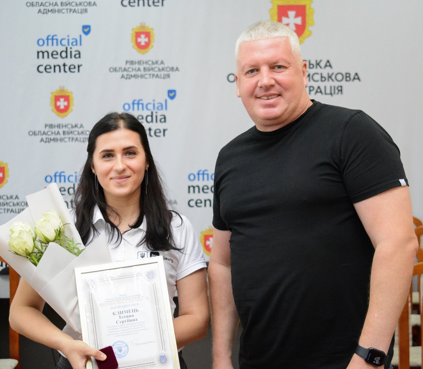 Жінка та чоловік позують для фото з грамотою та квітами. 