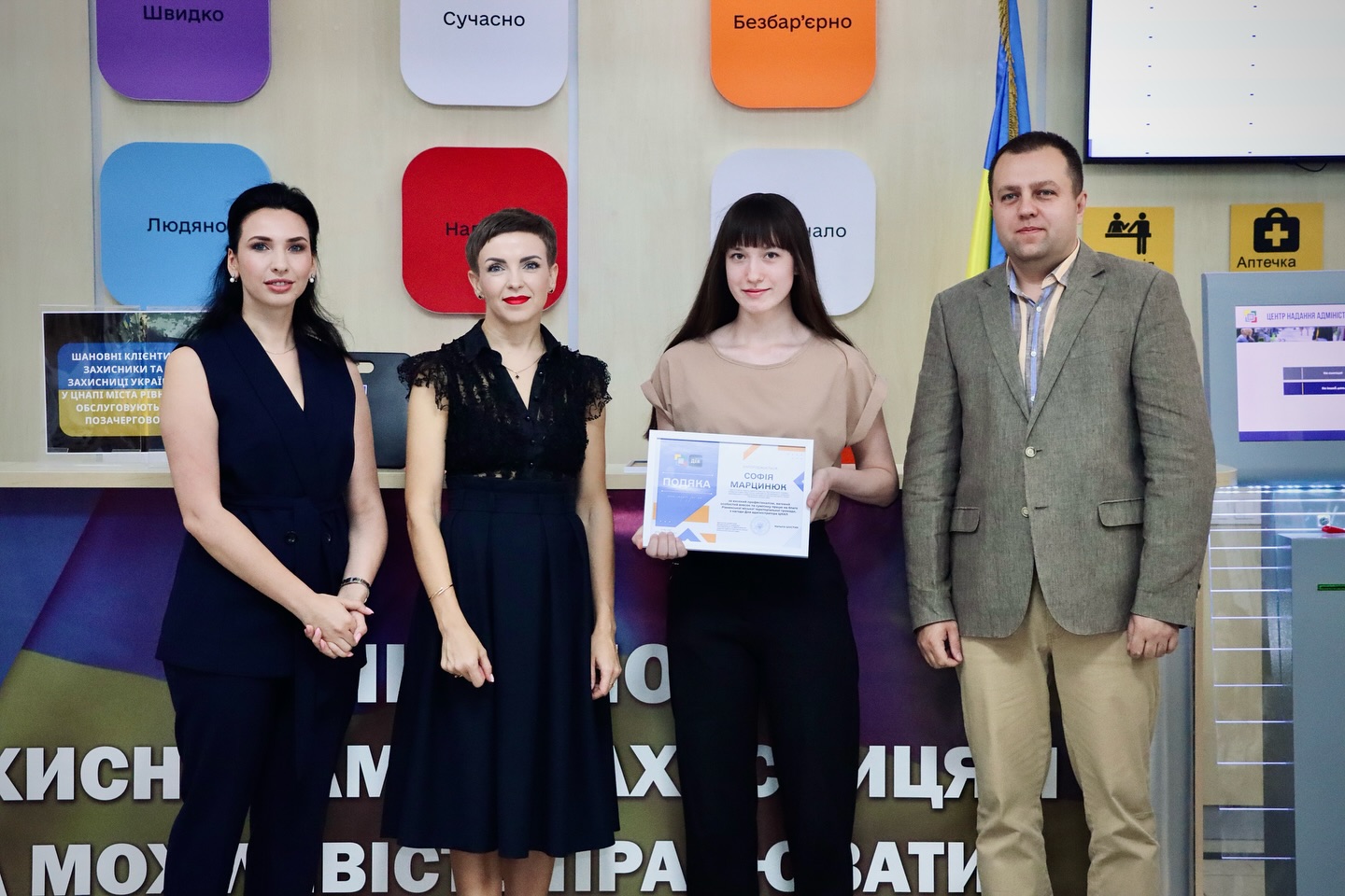 Три жінки з темним волоссям та чоловік позують для фото, одна жінка тримає подяку.