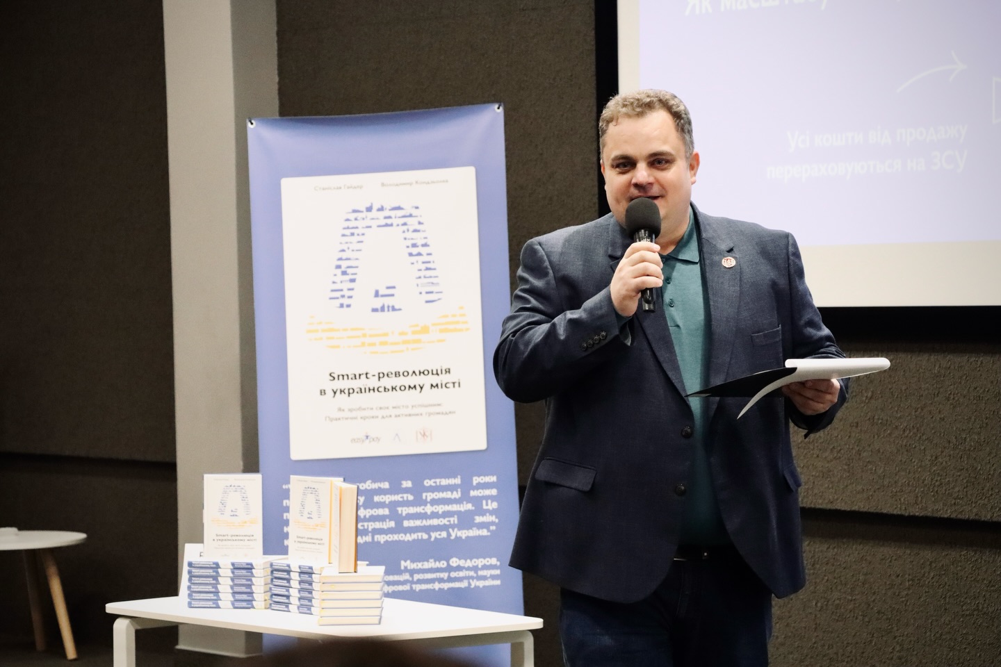 Чоловік у піджаку тримає у руці папку та говорить у мікрофон.