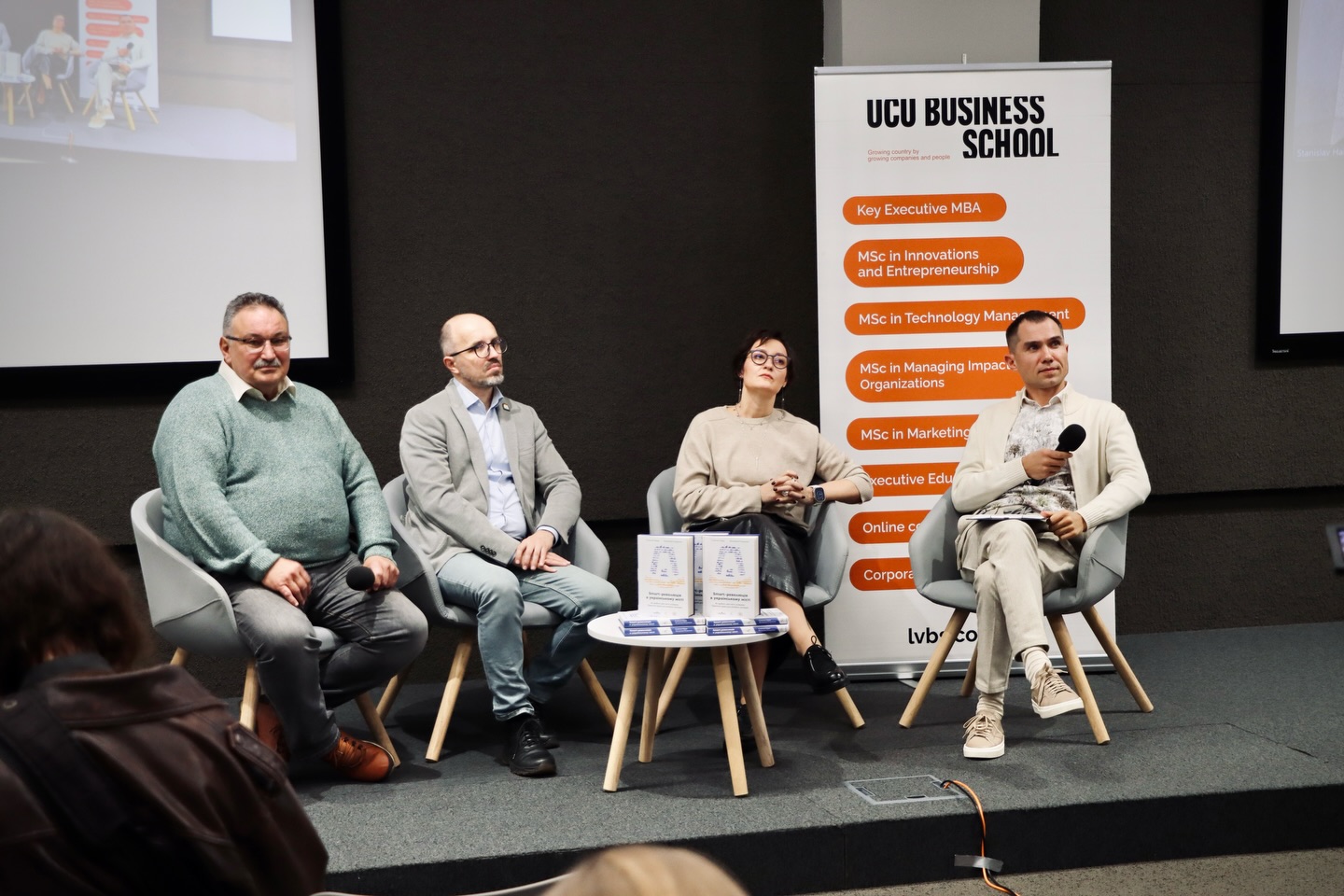 Жінка та троє чоловіків сидять перед екраном та банером.