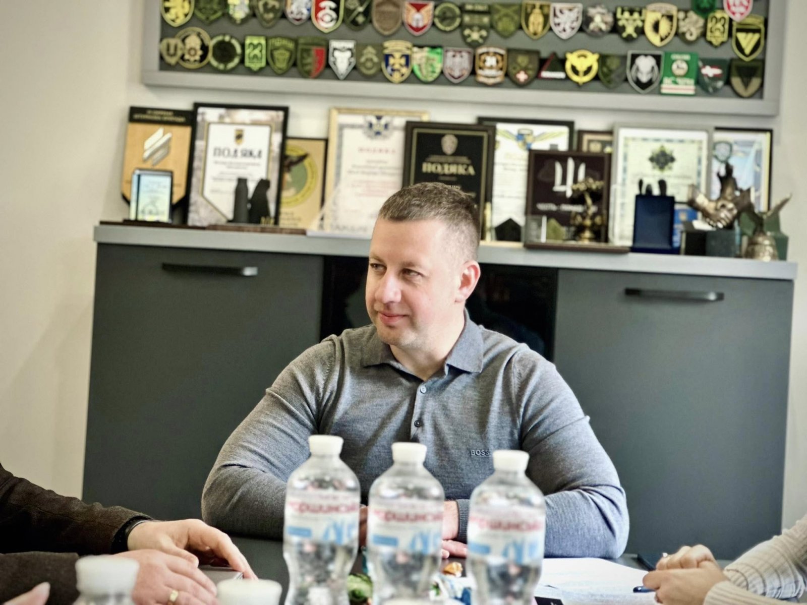 Чоловік у сірому светрі сидить за столом. За його спиною шафа нагородами, грамотами та емблемами.