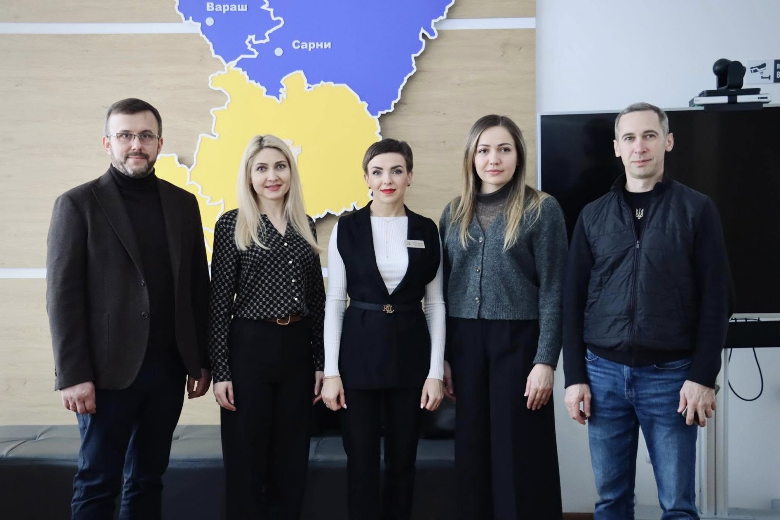 Група людей з двох чоловіків та трьох жінок стоять у приміщенні на фоні карти Рівненської області.