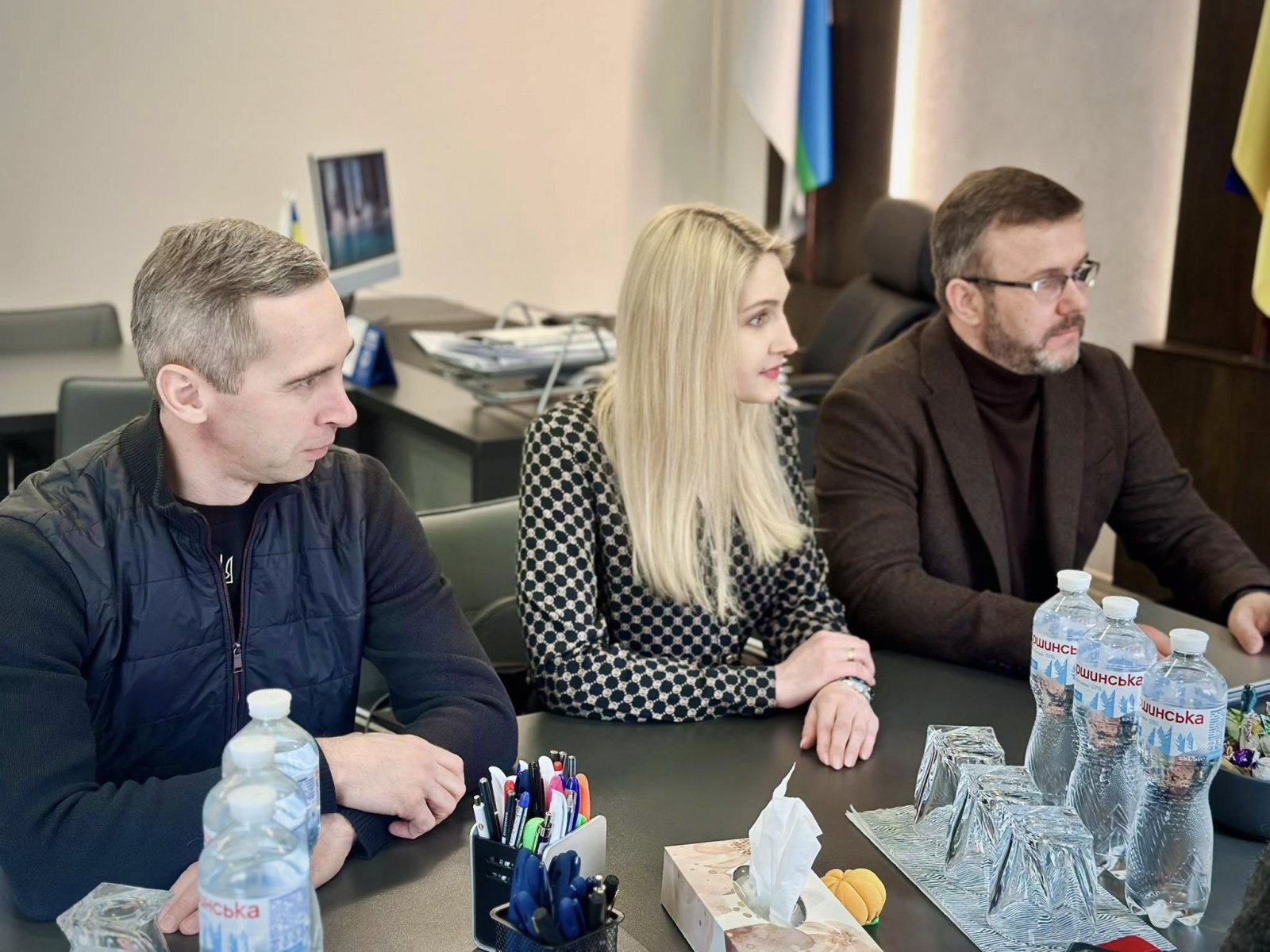 Жінка та двоє чоловіків сидять за столом. На столі стоїть вода та канцелярські приладдя.