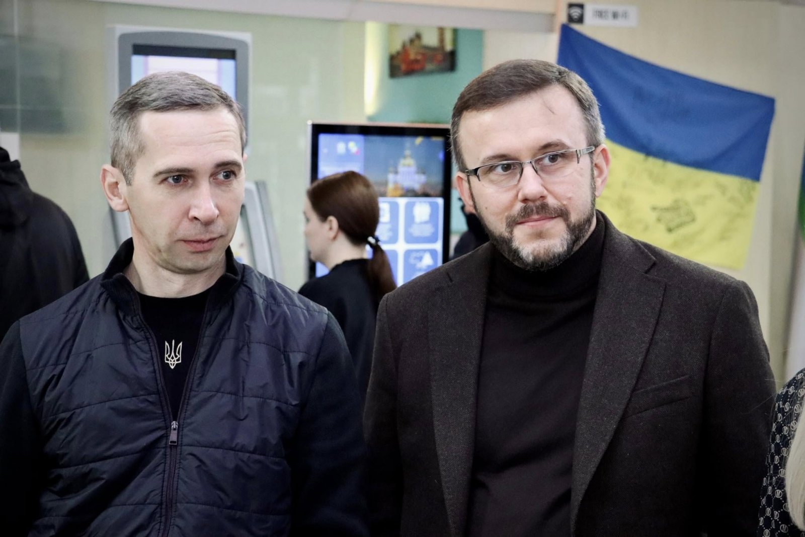 Два чоловіки стоять у приміщенні. На задньому фоні прапор України та інформаційний термінал.