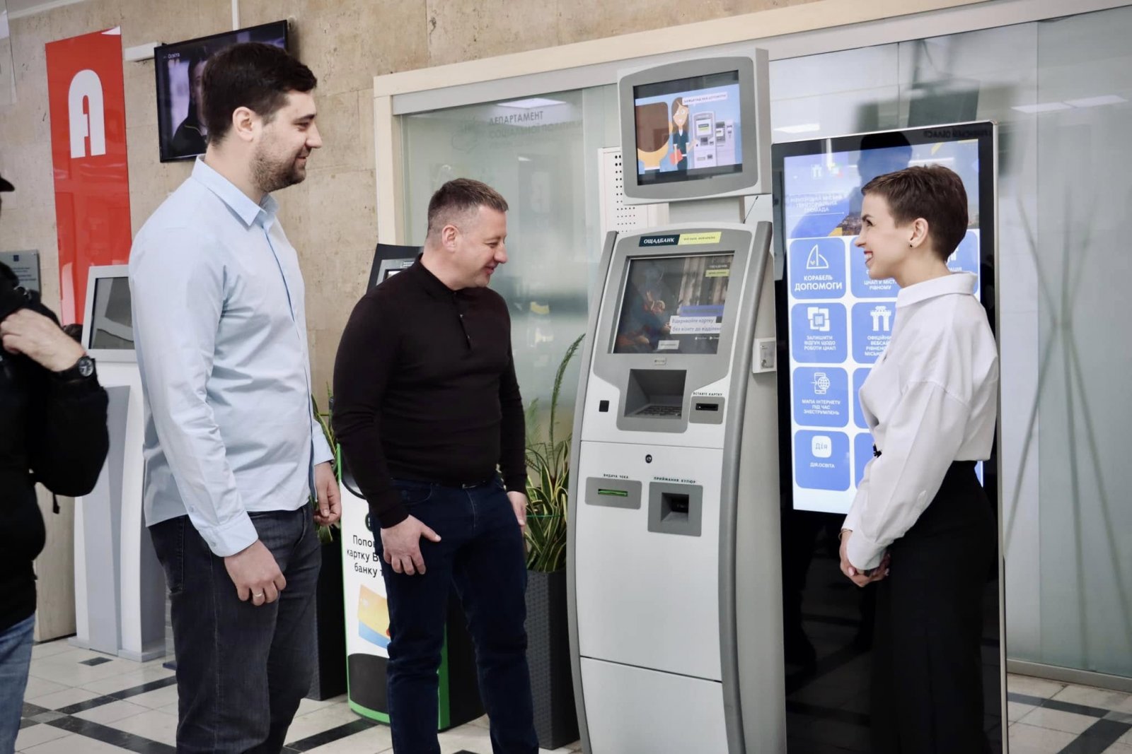 Двоє чоловіків спілкуються з жінкою біля інформаційного термінала та термінала банку.