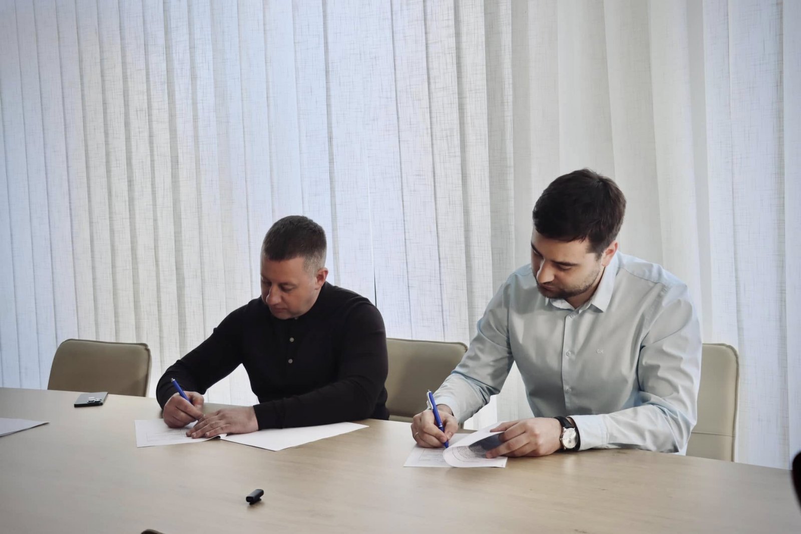 Двоє посадовців пишуть документи за столом. Фон із жалюзями, атмосфера офіційна.