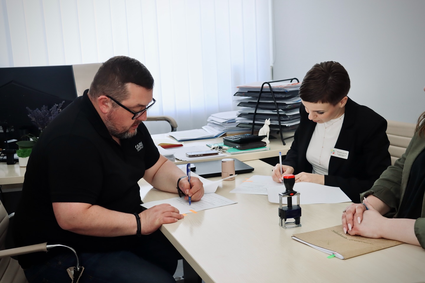 Чоловік і жінка підписують документи в офісі за столом із паперами та печаткою.