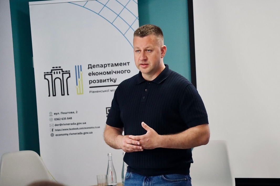Чоловік в темному одязі стоїть у приміщенні, на фоні - банер та два стільці