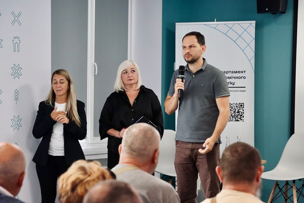 Чоловік говорить у мікрофон перед аудиторією, біля нього стоять дві жінки.