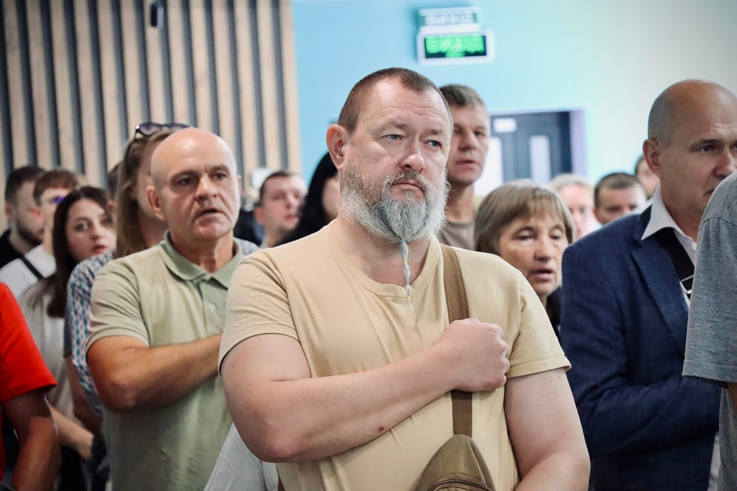 У приміщенні багато людей тримають руку на серці.