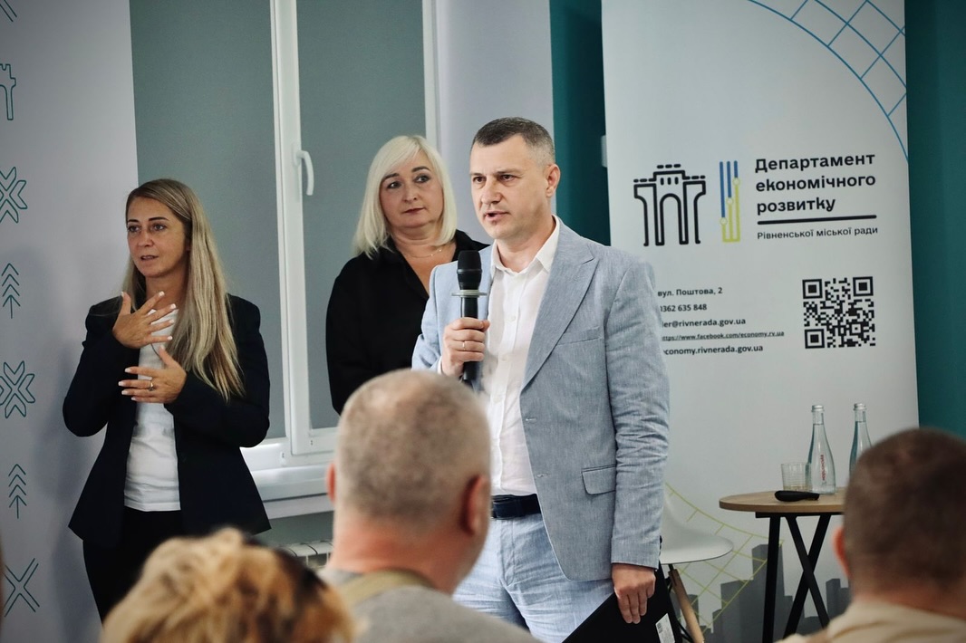 Чоловік у діловому одязі говорить у мікрофон перед аудиторією, позаду — дві жінки.