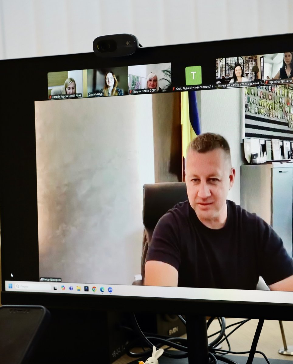 Онлайн-зустріч у Zoom, на екрані чоловік у темному одязі перед прапором України.