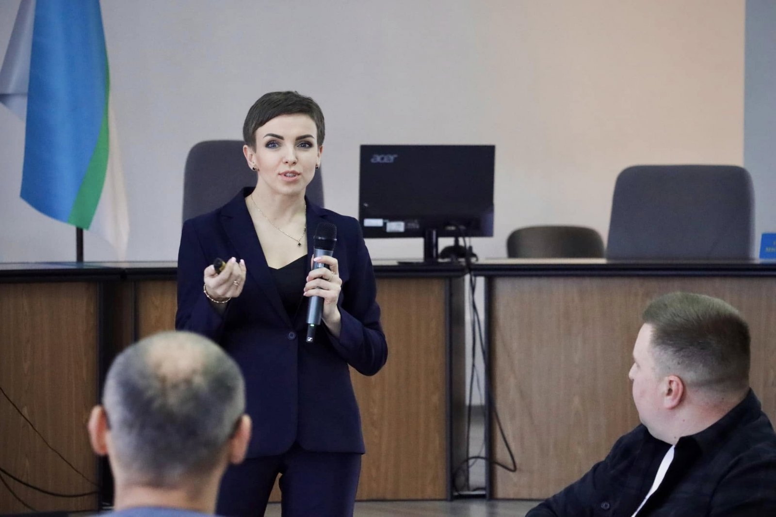 Жінка у темному костюмі з мікрофоном виступає перед аудиторією.